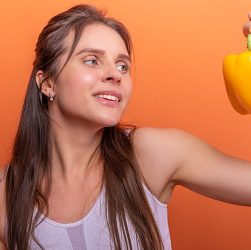 Woman in sports bra juggling choice between a donut and bell pepper on orange background.